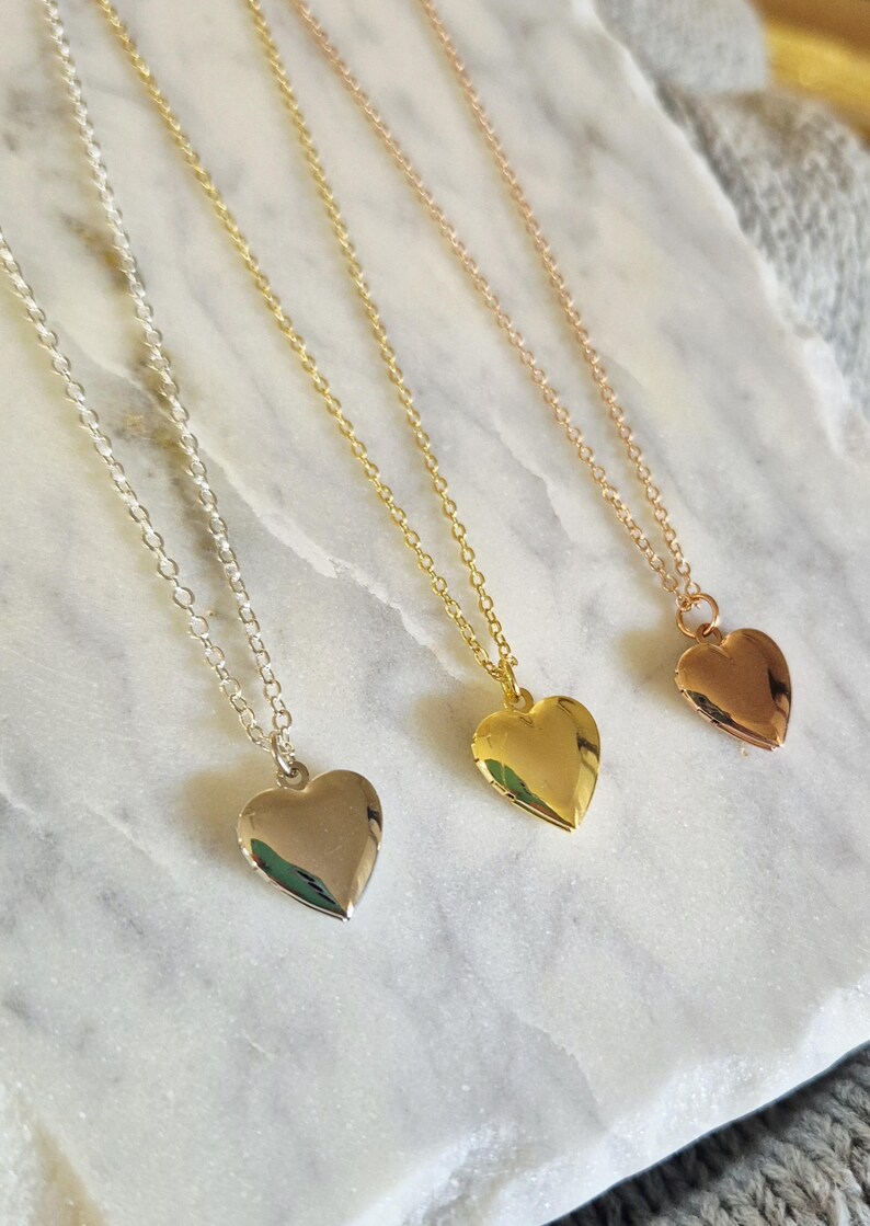 Puede incluir: Tres collares con colgantes en forma de coraz&oacute;n en plata, oro y oro rosa. Cada collar presenta una cadena delicada y un colgante de coraz&oacute;n liso y pulido. Los collares se exhiben sobre una superficie de m&aacute;rmol blanco y gris.