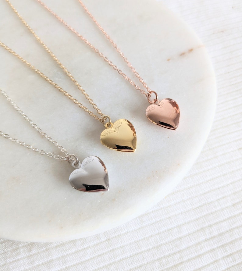 Puede incluir: Tres collares con colgantes en forma de coraz&oacute;n en plata, oro y oro rosa. Cada collar presenta una fina cadena y un colgante de coraz&oacute;n pulido. Los colgantes se exhiben sobre una superficie de m&aacute;rmol blanco, mostrando los diferentes acabados met&aacute;licos.