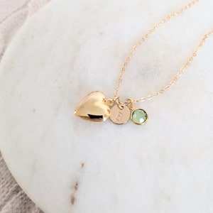 Puede incluir: Un collar dorado con un relicario en forma de coraz&oacute;n, un dije circular con la letra "B" y una gema verde. El collar se exhibe sobre una superficie de m&aacute;rmol blanco.