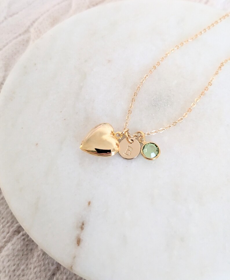 Puede incluir: Un collar dorado con un relicario en forma de coraz&oacute;n, un dije circular con la letra "B" y una gema verde. El collar se exhibe sobre una superficie de m&aacute;rmol blanco.