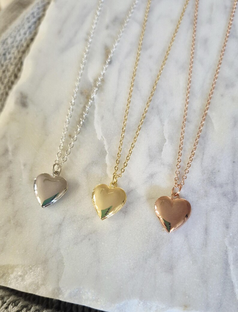 Puede incluir: Tres collares con colgantes en forma de coraz&oacute;n, plata, oro y oro rosa. Cada collar tiene una cadena delicada y un medall&oacute;n pulido en forma de coraz&oacute;n. Los colgantes se muestran sobre una superficie de m&aacute;rmol blanco.