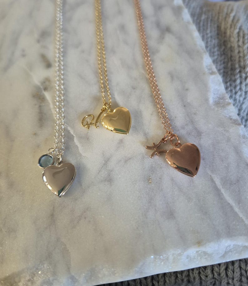 Puede incluir: Tres collares con colgantes en forma de coraz&oacute;n en plata, oro y oro rosa. El collar de plata tiene un dije de piedra preciosa azul. El collar de oro tiene un dije inicial. El collar de oro rosa tiene un dije de estrella.