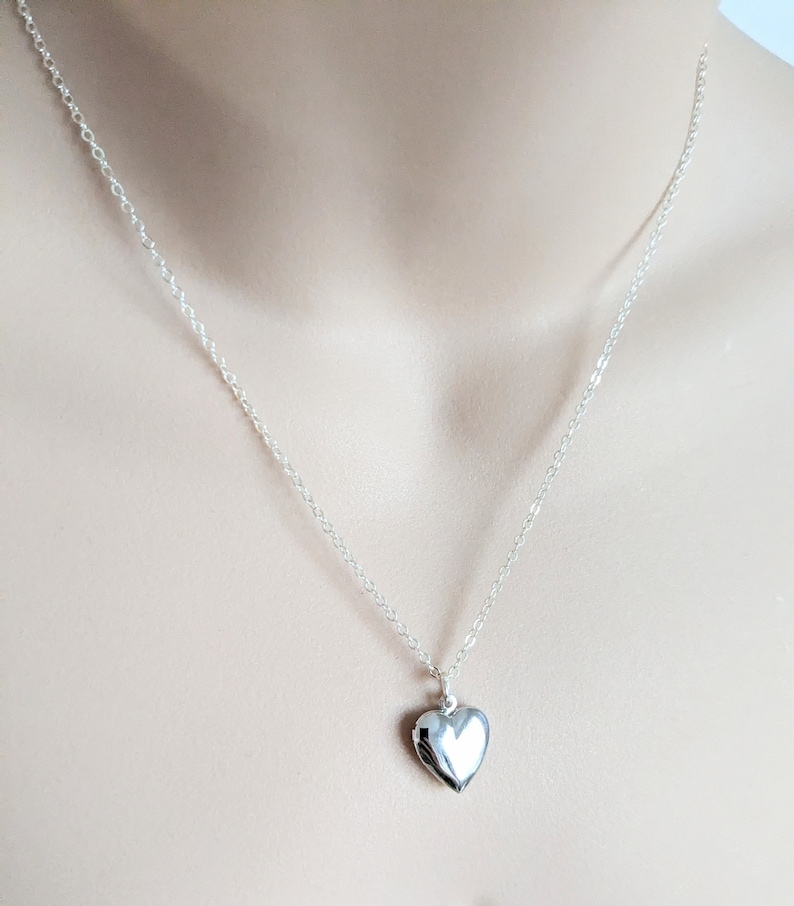 Puede incluir: Un collar con un colgante en forma de coraz&oacute;n plateado. El relicario cuelga de una fina cadena. El colgante en forma de coraz&oacute;n es liso y reflectante, con un dise&ntilde;o de bisagra. El collar se muestra sobre un fondo neutro.