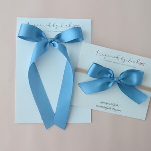 May include: Two light blue satin bows, one with long streamers, and a smaller bow on a headband. The bows are on white cards with the text "Inspired by Leah" and a website address. The background is a light pink.