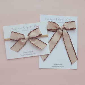 May include: Two beige hair bows with dark brown scalloped edges attached to white display cards labelled Inspired by Leah. One bow is on a thin elastic headband, while the other is a long-tailed bow. Both cards feature the text @inspiredbyleah.