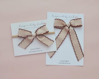 Op de afbeelding: Twee beige haarstrikken met donkerbruine geschulpte randen op witte displaykaarten met het label Inspired by Leah. Eén strik zit op een dunne elastische haarband, de andere heeft lange linten. Beide kaarten bevatten de tekst @inspiredbyleah.