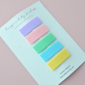May include: Five pastel-colored rectangular hair clips with a scalloped edge are arranged diagonally on a white card. The colors are lavender, pink, mint green, light blue, and yellow. The card has text that reads "Inspired by Leah" and "Handmade Hair Accessories for Girls."