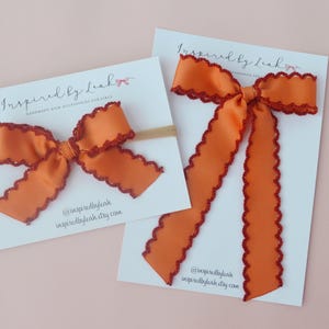 Könnte beinhalten: Zwei orangefarbene Haarschleifen mit dunkelroten, gewellten Rändern auf weißen Präsentationskarten mit der Aufschrift Inspired by Leah. Eine Schleife ist an einem dünnen, beigen elastischen Haarband befestigt, die andere hat lange Bänder.