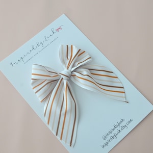 Op de afbeelding: Een witte haarstrik met verticale strepen in bruin- en beigetinten. De strik is in het midden geknoopt en heeft lange, vloeiende uiteinden. De strik ligt op een witte kaart met de tekst "Inspired by Leah" en "Handmade Hair Accessories for Girls".