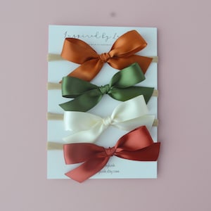 May include: Set of four hair bows in rust, green, ivory, and burgundy colors. The bows are made of satin ribbon and are attached to nude elastic headbands. The headbands are designed for babies and toddlers.