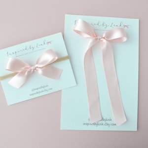 Puede incluir: Dos lazos de pelo de satén rosa, uno en una diadema elástica de color nude y el otro con colas largas. Los lazos se muestran en tarjetas blancas con el texto "Inspired by Leah" y la dirección del sitio web "inspiredbyleah.etsy.com".