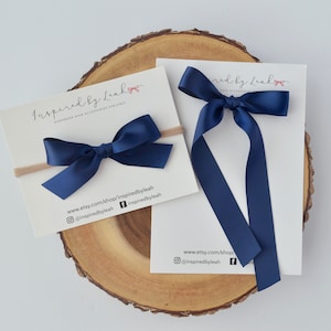 May include: Navy blue satin ribbon hair bow on a nude elastic headband. The bow is tied in a simple knot and the headband is made of a soft, stretchy material. The bow is perfect for adding a touch of elegance to any outfit.