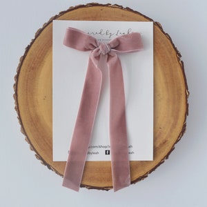 Puede incluir: Un lazo para el pelo de terciopelo rosa polvoriento con colas largas. El lazo está atado en un nudo simple y se muestra sobre una superficie de madera. El texto "Inspired by Leah" es visible en la tarjeta detrás del lazo.