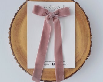 May include: A dusty rose velvet hair bow with two long, trailing ribbon tails. The bow is attached to a white rectangular display card featuring the text Inspired by Leah and social media icons, centered on a circular natural wood slice.