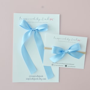 Könnte beinhalten: Zwei hellblaue Satin-Haarbänder auf weißen Karten mit dem Text "Inspired by Leah Handmade Hair Accessories for Girls" und Website-Informationen. Das kleinere Band ist auf einem nudefarbenen elastischen Haarreif.