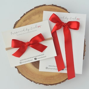 May include: Two red satin hair bows, one on a nude elastic headband and the other with long tails. The bows are handmade and are displayed on a wooden slice with a business card that reads "Inspired by Leah Handmade Hair Accessories for Girls" and includes social media handles.