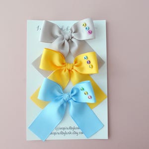May include: Three colourful hair bows in grey, yellow, and blue are displayed on a white card. Each bow has a small embellishment with the letters "A, B, C". The bows are arranged on a pink background.