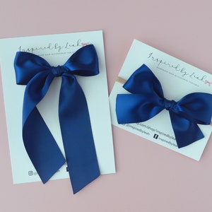 Puede incluir: Dos lazos para el pelo de satén azul marino, uno grande y otro pequeño. El lazo más grande tiene colas largas y está en una tarjeta blanca con el texto "Inspired by Leah Handmade Hair Accessories for Girls". El lazo más pequeño está en una tarjeta blanca con el texto "Inspired by Leah Handmade Hair Accessories for Girls" y la dirección del sitio web "inspiredbyLeah.com/shop/inspiredbyLeah @inspiredbyLeah f InspiredbyLeah".