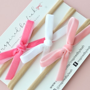 May include: Three velvet hair bows in pink, white, and light pink, tied with a knot, on nude elastic headbands. The headbands are displayed on a white card with the text "Inspired by Leah" and social media handles.