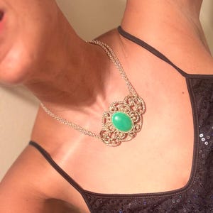 May include: A silver-toned necklace featuring an oval, light green gemstone set in an ornate, filigree-style pendant. The necklace has a double chain and is worn against a black sequined top.