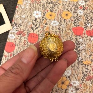 May include: A gold-colored, textured pendant with a small chain. The pendant is round with a detailed surface. The background features a floral pattern with flowers in shades of yellow, red, and white on a beige background.