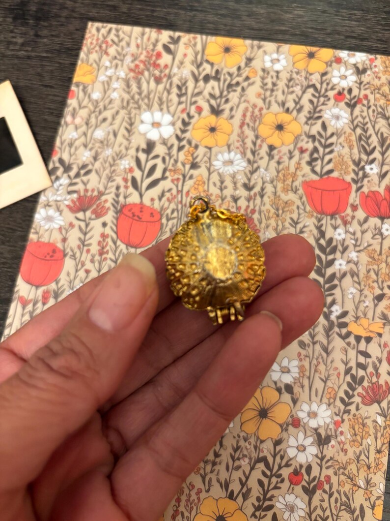 May include: A gold-colored, textured pendant with a small chain. The pendant is round with a detailed surface. The background features a floral pattern with flowers in shades of yellow, red, and white on a beige background.