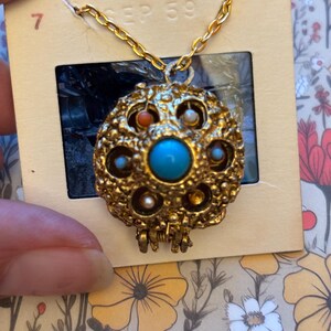 May include: A gold-toned pendant necklace with a round, textured charm. The charm features a large turquoise stone in the center, surrounded by smaller stones in various colors. The necklace is attached to a gold chain. The slide mount reads "MADE IN U.S.A." and "SEP 59".