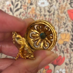 May include: Ornate gold-colored jewelry piece with intricate detailing. The piece features a central turquoise-colored stone and a decorative, openwork design. A small, dangling element is attached by a delicate chain. The background is a floral pattern.