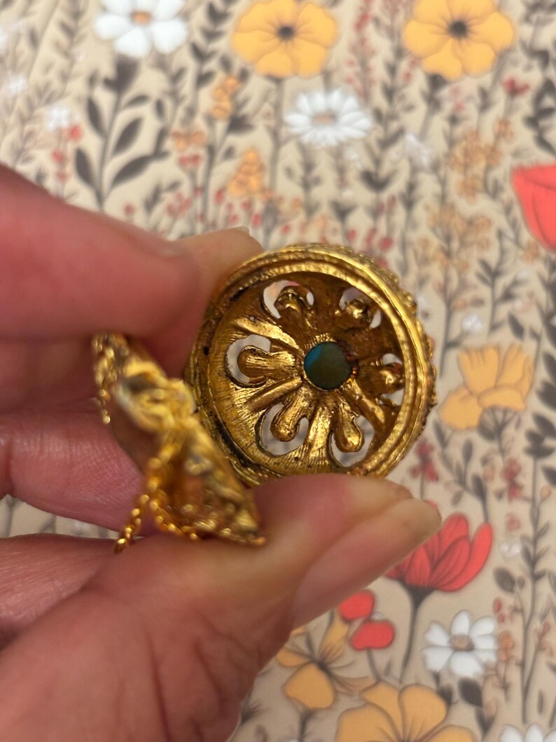 May include: Ornate gold-colored jewelry piece with intricate detailing. The piece features a central turquoise-colored stone and a decorative, openwork design. A small, dangling element is attached by a delicate chain. The background is a floral pattern.