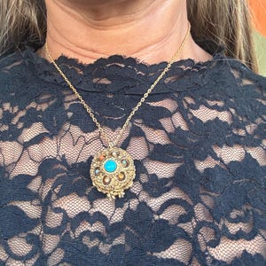 May include: Gold-toned pendant necklace with a round, ornate pendant. The pendant features a central turquoise stone surrounded by smaller stones and intricate metalwork. The necklace is worn over a black lace top.