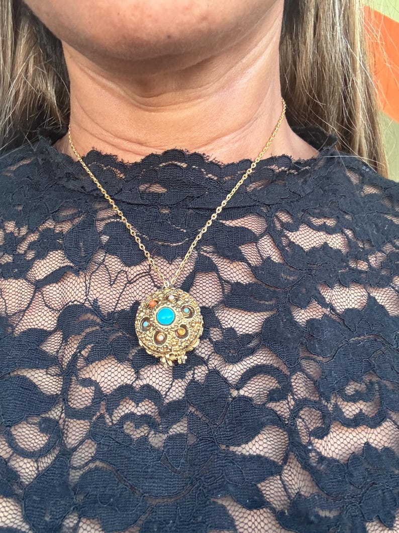 May include: Gold-toned pendant necklace with a round, ornate pendant. The pendant features a central turquoise stone surrounded by smaller stones and intricate metalwork. The necklace is worn over a black lace top.