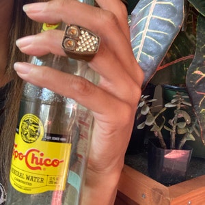 May include: A rectangular ring with a bronze-colored band and a face covered in small, sparkling crystals and two circular amber-colored accents. The ring is held near a clear glass bottle of Topo Chico mineral water.