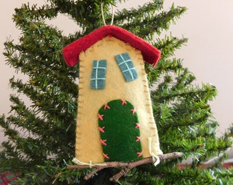 May include: A felt house ornament with a red roof, yellow walls, green door, and blue windows. The ornament is hanging from a branch of a Christmas tree.