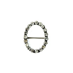 May include: A silver oval-shaped brooch with a row of clear rhinestones around the edge.