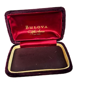 May include: An empty, burgundy velvet jewelry box with gold trim. The inside of the box is lined with red satin and has the words "Bulova Fifth Avenue New York" printed in gold.