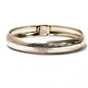 May include: A polished sterling silver hinged bangle bracelet featuring an engraved floral pattern along the outer surface. The interior clasp mechanism is clearly stamped with the text STERLING SILVER, indicating the metal composition.
