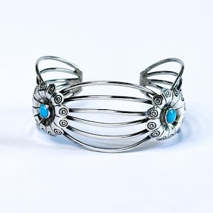 May include: A silver cuff bracelet with a floral design and two turquoise-colored stones. The bracelet has a series of parallel bands and decorative floral elements on each end. The bracelet is displayed on a white background.