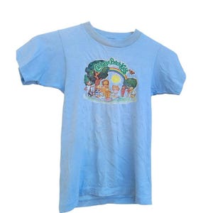 May include: Light blue t-shirt featuring a colorful graphic of Cabbage Patch Kids under a rainbow. The graphic includes trees, a sun, and several Cabbage Patch Kids characters. The shirt has short sleeves and a crew neck.