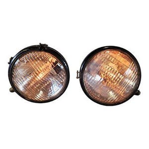 May include: Two vintage-style round headlights with black rims. The glass lenses have a textured pattern and a warm, amber glow. The headlights are designed for a classic or custom vehicle.