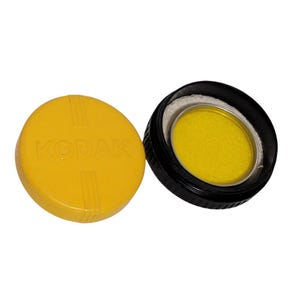 May include: A yellow round container with the word "KODAK" printed on the lid. The container holds a yellow filter.