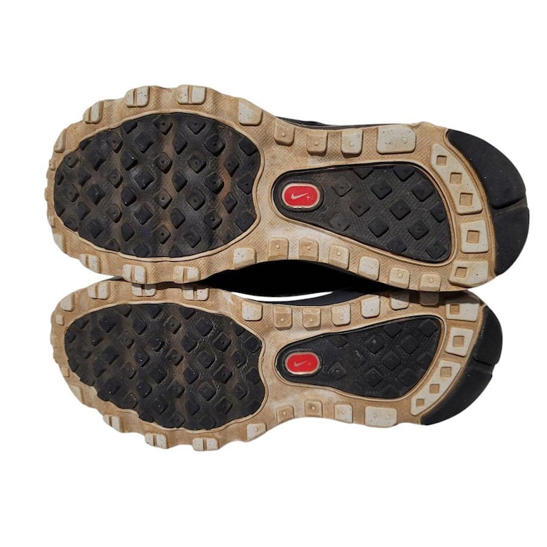 May include: A pair of black athletic shoes, viewed from the soles. The soles are black with a textured pattern and a red Nike logo. The outer edges are tan and have a lugged design for traction.