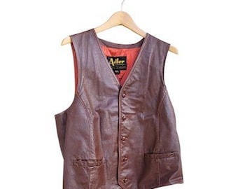 May include: A brown leather vest with a V-neck and button closure, hanging on a wooden hanger. The vest has two front pockets and a red interior lining. The label reads "Adler".