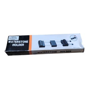 May include: A rectangular box with the words "WATERSTONE HOLDER" in bold white letters on a black background. The box features a diagram of a waterstone holder with a black and silver design. An orange stripe runs along the top.