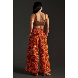 K&ouml;nnte beinhalten: Ein braunes Crop-Top und eine Hose mit weitem Bein und einem lebendigen Blumenmuster. Die Hose ist haupts&auml;chlich orange mit roten und schwarzen Blumendesigns. Das Outfit ist l&auml;ssig und stilvoll.