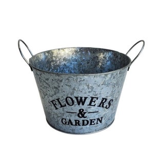 May include: A galvanized metal bucket with two arched handles. The bucket has a tapered shape and features the words "FLOWERS & GARDEN" in black lettering. The bucket is designed for gardening or decorative use.