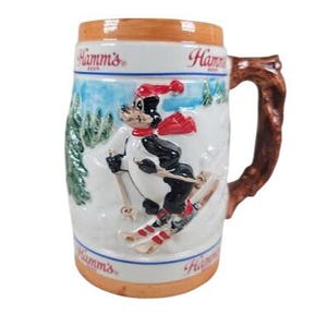 May include: Vintage Hamm's Beer ceramic stein. The stein features a cartoon skier in a red hat and scarf, skiing down a snowy hill. The handle is designed to look like a tree branch. The stein has the Hamm's Beer logo.