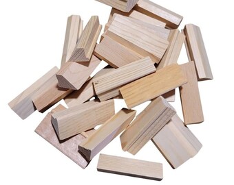 May include: A pile of unfinished wooden blocks, varying in size and shape, are scattered against a white background. The blocks are made of light-colored wood, with visible grain patterns. Some blocks are rectangular, while others have angled edges, suggesting a building or stacking toy.