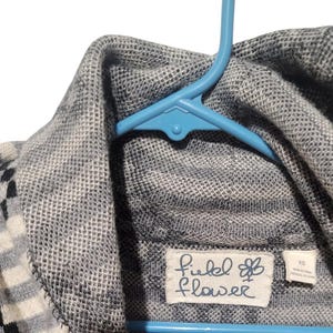 May include: A gray and white striped sweater with a black and white pattern. The sweater has a turtleneck collar and a label that reads "Field of Flower" with a small flower graphic.