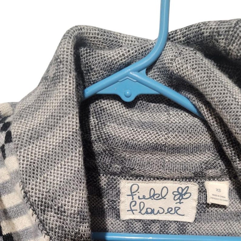 May include: A gray and white striped sweater with a black and white pattern. The sweater has a turtleneck collar and a label that reads "Field of Flower" with a small flower graphic.