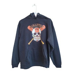 May include: A navy blue hoodie with a front graphic of a skull wearing a baseball helmet and crossed bats. The word "GIGANTES" is above the skull in orange lettering. The hoodie has a front pocket and a drawstring hood.